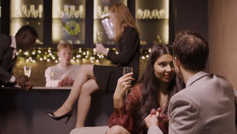 group of friends celebrating the new year's party, a couple are talking sitting at a table, while the others are at the bar eating grapes and drinking champagne