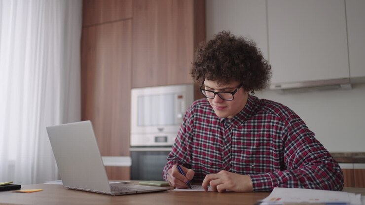 En attraktiv, krølhåret mandlig studerende med briller studerer hjemme. Han bruger sin bærbare computer til at skrive og tage noter i en notesbog. Den studerende ser et online fjernundervisningsseminar på sin bærbare computer.