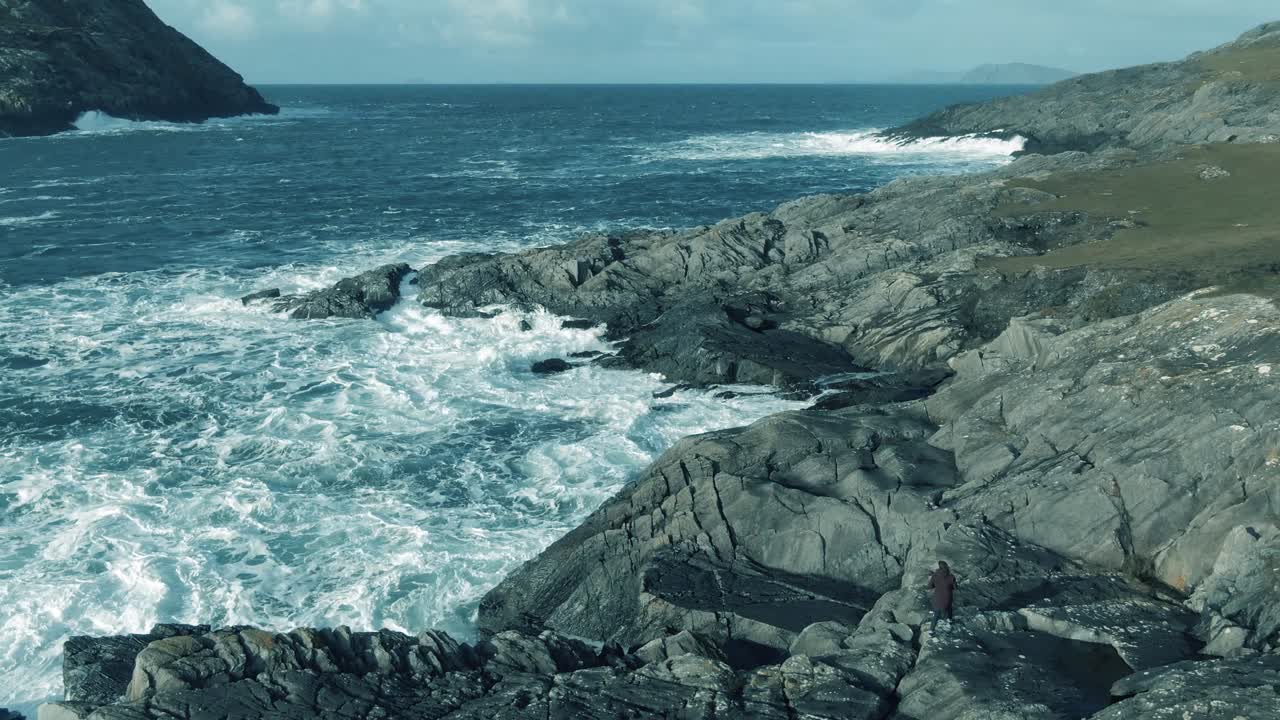 Slow aerial pan out of irish rocky shore line with waves crashing on the shores and spray in the air with a photographer on the shoreline