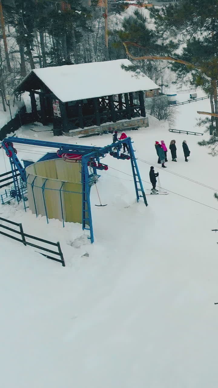 skiers stand on hill top near lift station with rope tow and dark wooden arbor in snowy pine tree forest aerial view