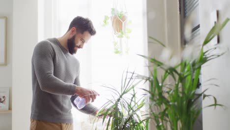 Video-of-happy-caucasian-man-watering-houseplants-at-home,-misting-with-spray,-copy-space