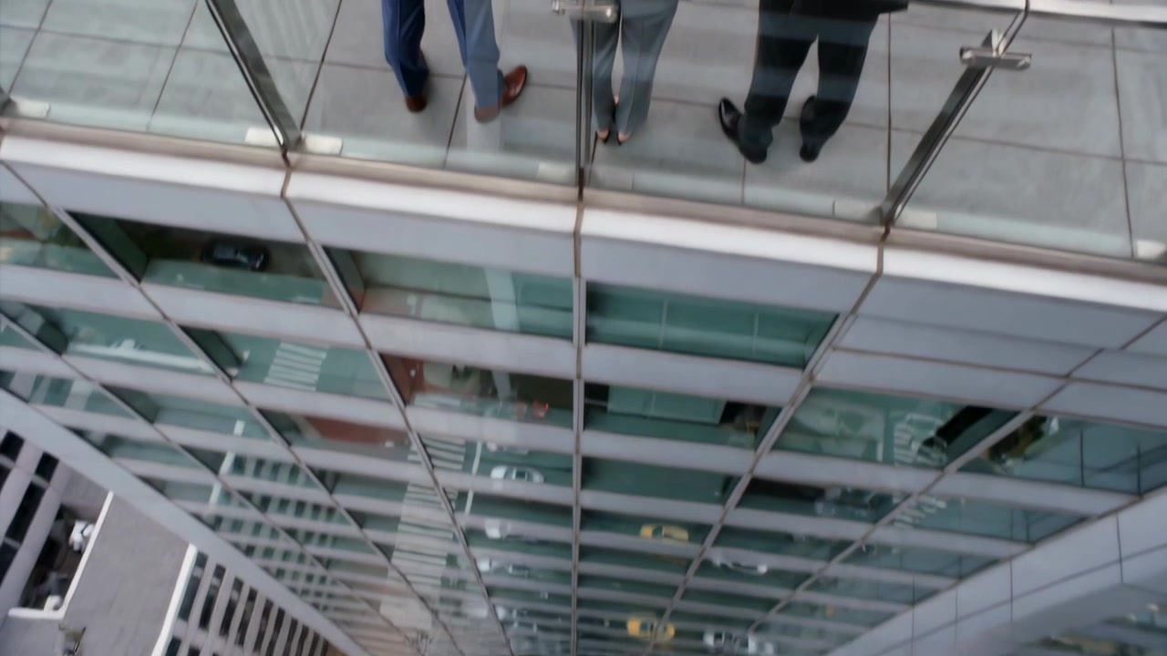 Câmera inclinando, revelando a parede do escritório, enquanto três homens de terno estão no corrimão da varanda observando o tráfego.