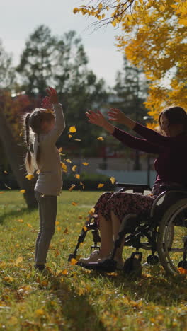 woman with paralysis plays with daughter in park. woman throws up dry leaves having fun with little girl. active rest during rehabilitation process