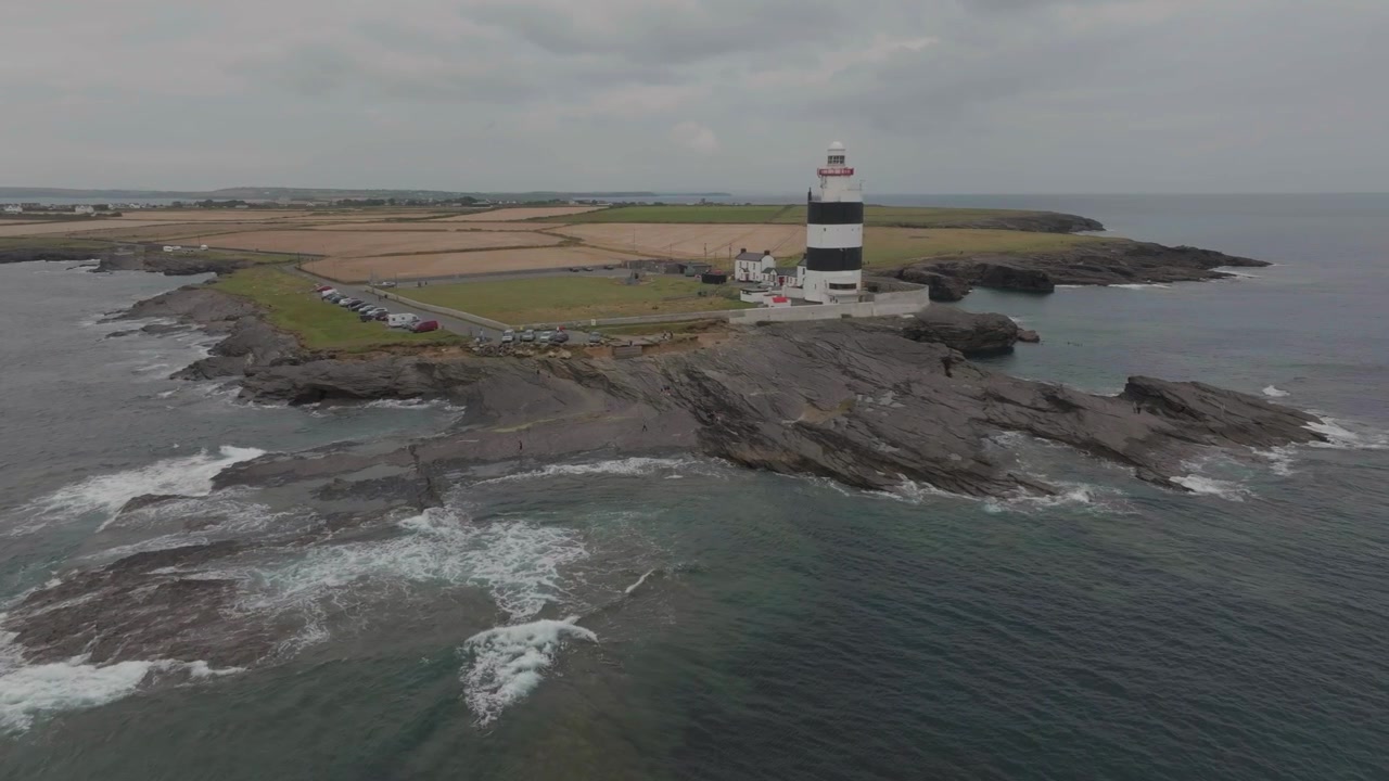 Et luftfoto af det historiske sort- og hvidstribede Hook Fyr, beliggende på en barsk klippefyldt halvø i County Wexford, Irland, under en overskyet himmel.