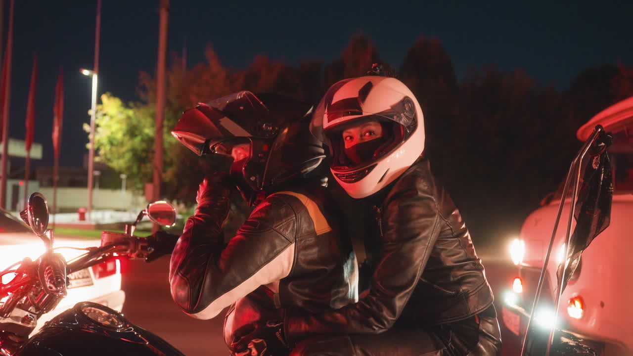Lady lifts visor smiling and making playful gesture while seated behind her brother on motorcycle, both in helmets and leather jackets, waiting at traffic stop under city night lights