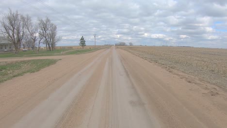 point of view driving on a straight stretch of gravel road, past a farm yard, a crossroad and harvested fields