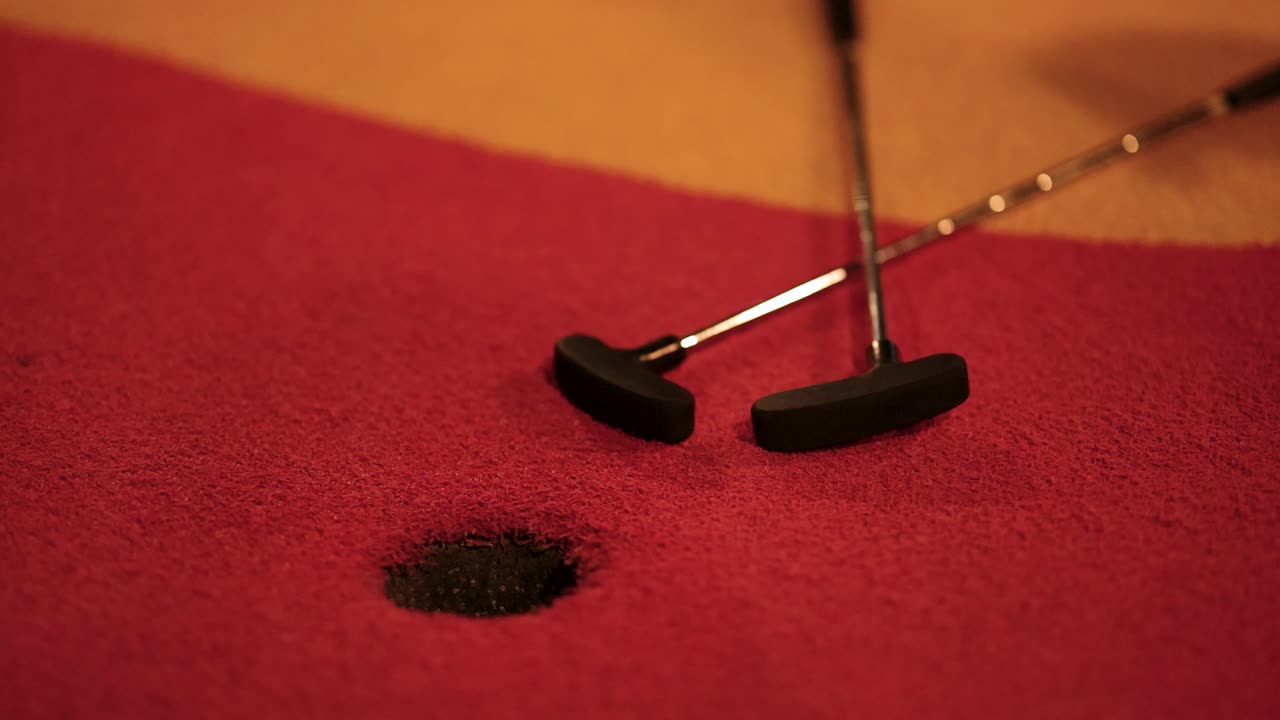 A putter hits an orange ball into a hole on a red mini-golf course under warm lighting