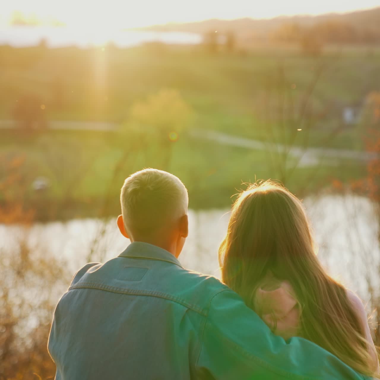 Young people sitting on grass and looking at sunset near the river. Guy embracing his girlfriend against autumn landscape in the evening