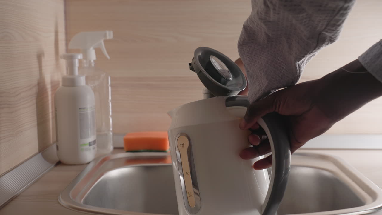 Close-up of man pouring water from white electric kettle into sink. Background features cleaning supplies, including a spray bottle and sponge, in a neat, minimalist kitchen setting. Soft morning light illuminates