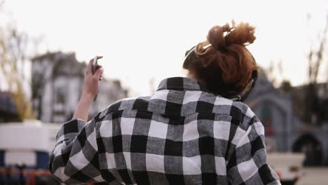 Backside,-slow-motion-shooting-of-a-girl-in-expressive-dance,-wearing-headphones.-Emotionally-whirling-her-head.-In-a-plaid-shirt-and-mobile-phone-in-her-hand