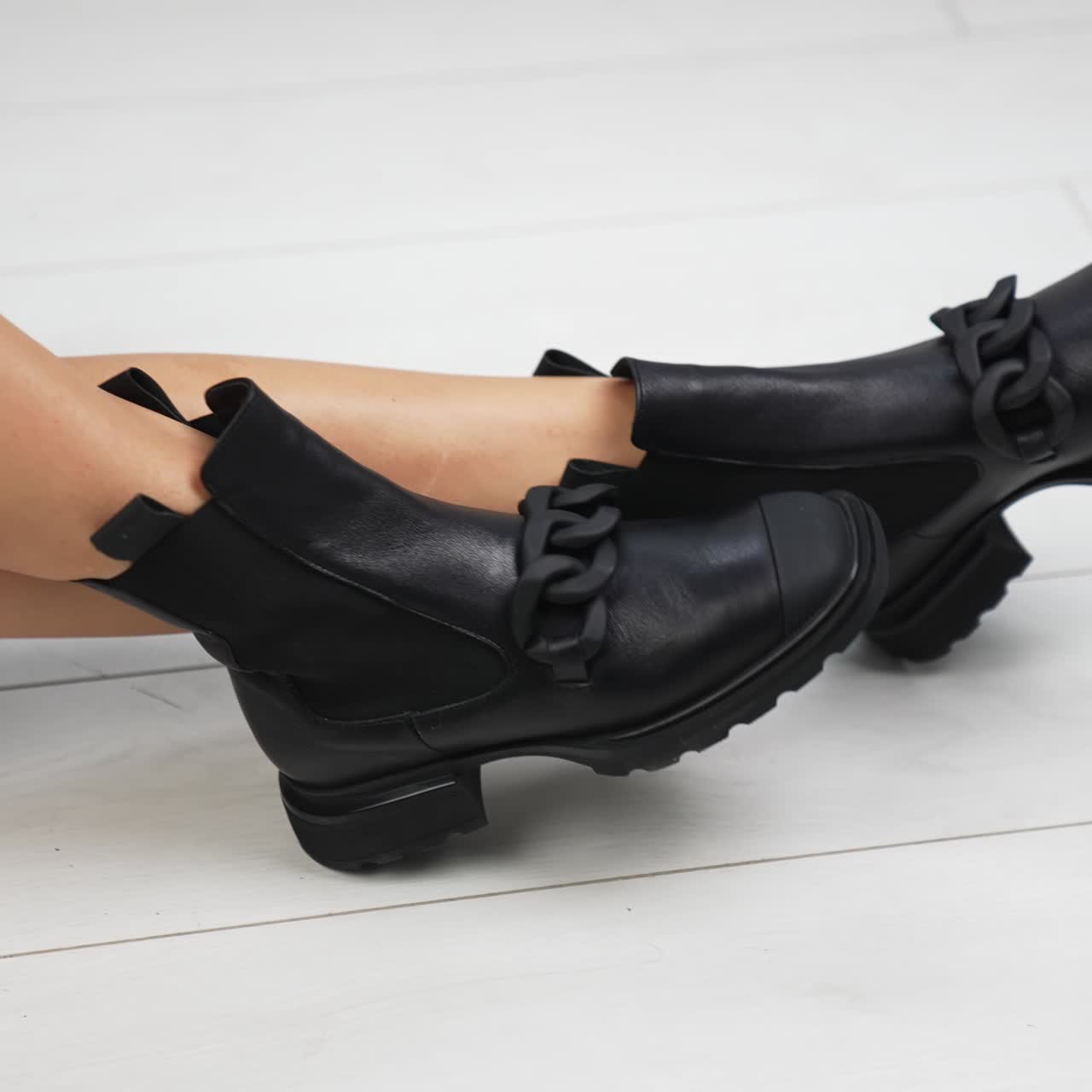 Bare-legged female sits on the floor in black boots. Lady touching the footwear with chains decorations on top. White floor