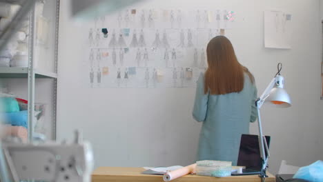 young fashion designer standing with back to camera looking at drawings sketches hanging on wall. woman is thinking about new trendy clothing collection.