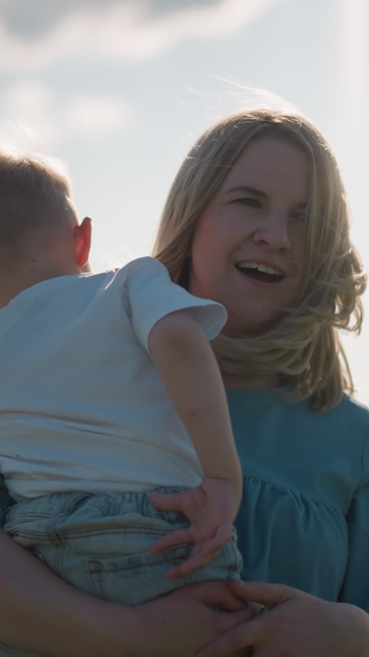A woman in a blue dress holds her young son, who is wearing a white top, as they both gaze out towards a serene lake. The peaceful moment captures the bond between mother and child