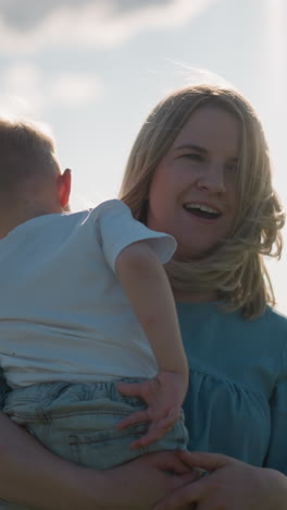a woman in a blue dress holds her young son, who is wearing a white top, as they both gaze out towards a serene lake. the peaceful moment captures the bond between mother and child