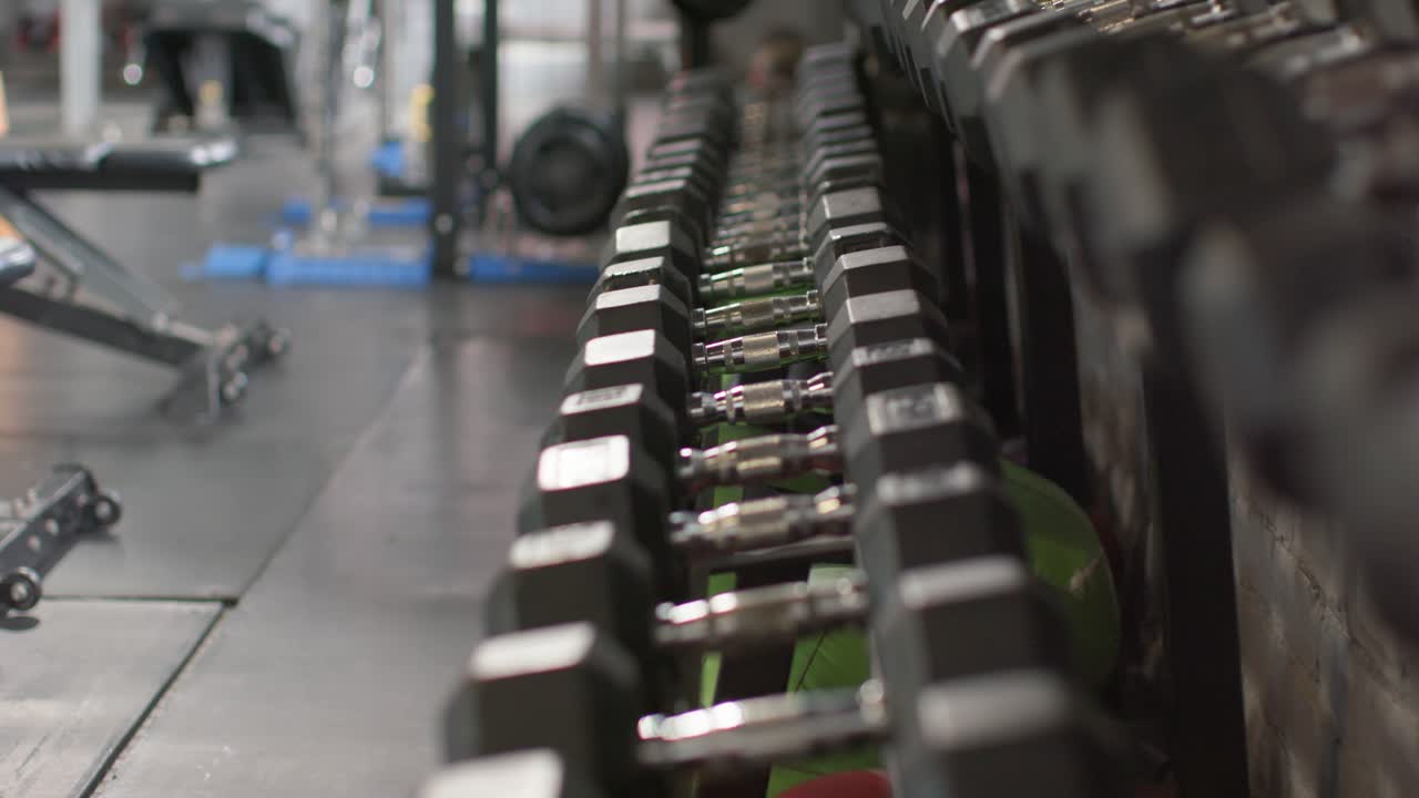 Camera is rolling forward and refocusing to reveal hex dumbbell rack with medicine balls in gym