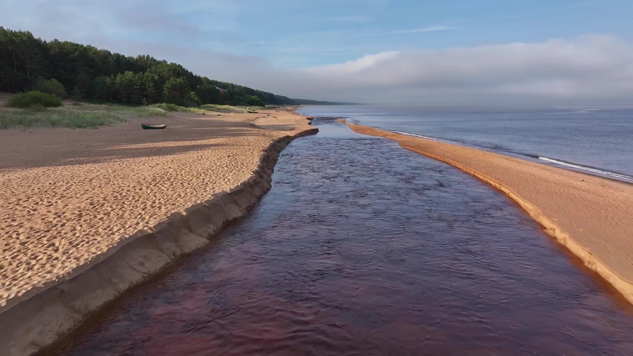 Riprese con drone a bassa quota che volano sopra il fiume Peterupe mentre sfocia nel Mar Baltico a Saulkrasti, Lettonia. Vista panoramica all'alba con spiagge sabbiose, mare calmo, foresta costiera e la naturale r...