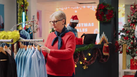 senior woman browsing through blazer racks in christmas decorated clothing store while husband pays at shop counter in background. clients making xmas purchases during winter holiday season