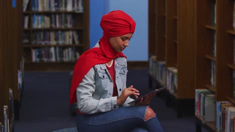 una estudiante asiática con un hijab rojo estudiando en una biblioteca y usando una tableta