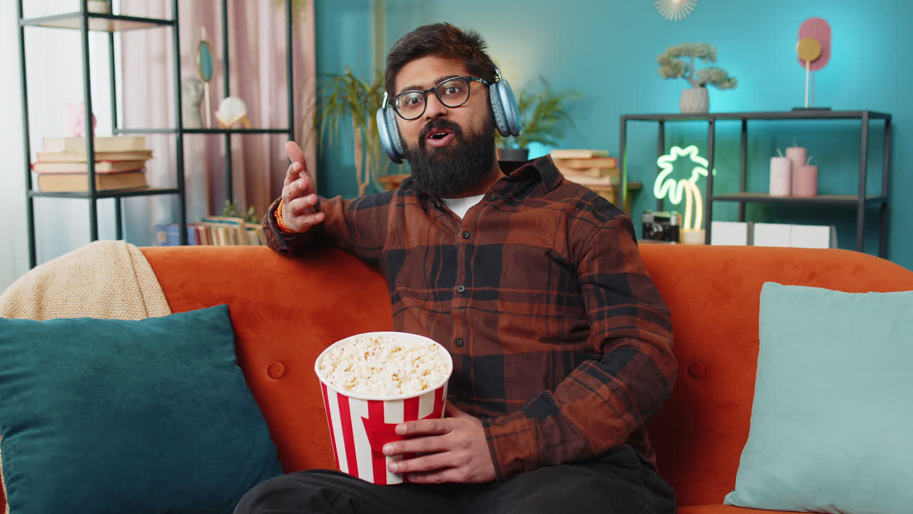 Relaxed indian man eating popcorn watching movie enjoying film during weekend sitting on home sofa