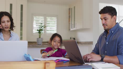 Hispanische-Eltern-Sitzen-Am-Tisch-Und-Arbeiten-Mit-Laptops-Und-Ihrer-Tochter-Zeichnen