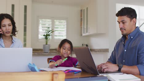 hispanic parents sitting at table working with laptops and daughter drawing