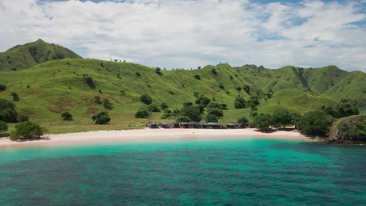A white sandy beach stretches along clear turquoise waters. Behind the beach, lush green hills rise, forming a natural backdrop under a bright tropical sky. Komodo Island, Indonesia.