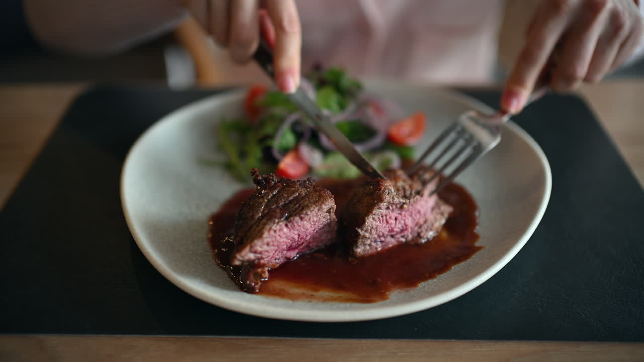 Close up of woman cutting juicy grilled steak served with rich sauce and fresh salad