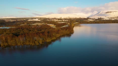 Vista-Aérea-Sobre-El-Lago-Lomond-Hacia-El-Campo-De-Golf-Cameron-Club,-Colinas-Nevadas-Al-Fondo-Sobre-El-Lago-En-Calma-Que-Reflejan-El-Cielo-Azul-Arriba