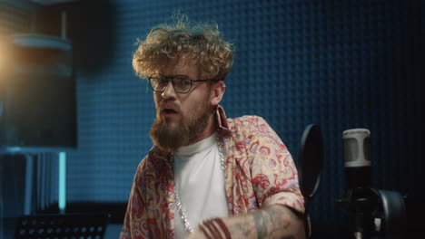 joven y elegante cantante hipster con anteojos y cabello rizado que mira a la cámara alegremente y lanza besos mientras está parado en el micrófono en el estudio de sonido