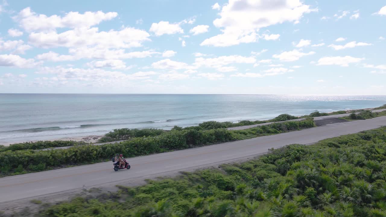 멕시코 열대 섬 코수멜에 있는 태평양 해안을 따라 고속도로를 따라 빨간 스쿠터를 운전하는 젊은 부부의 궤도 드론 샷이 4k로 촬영되었습니다.