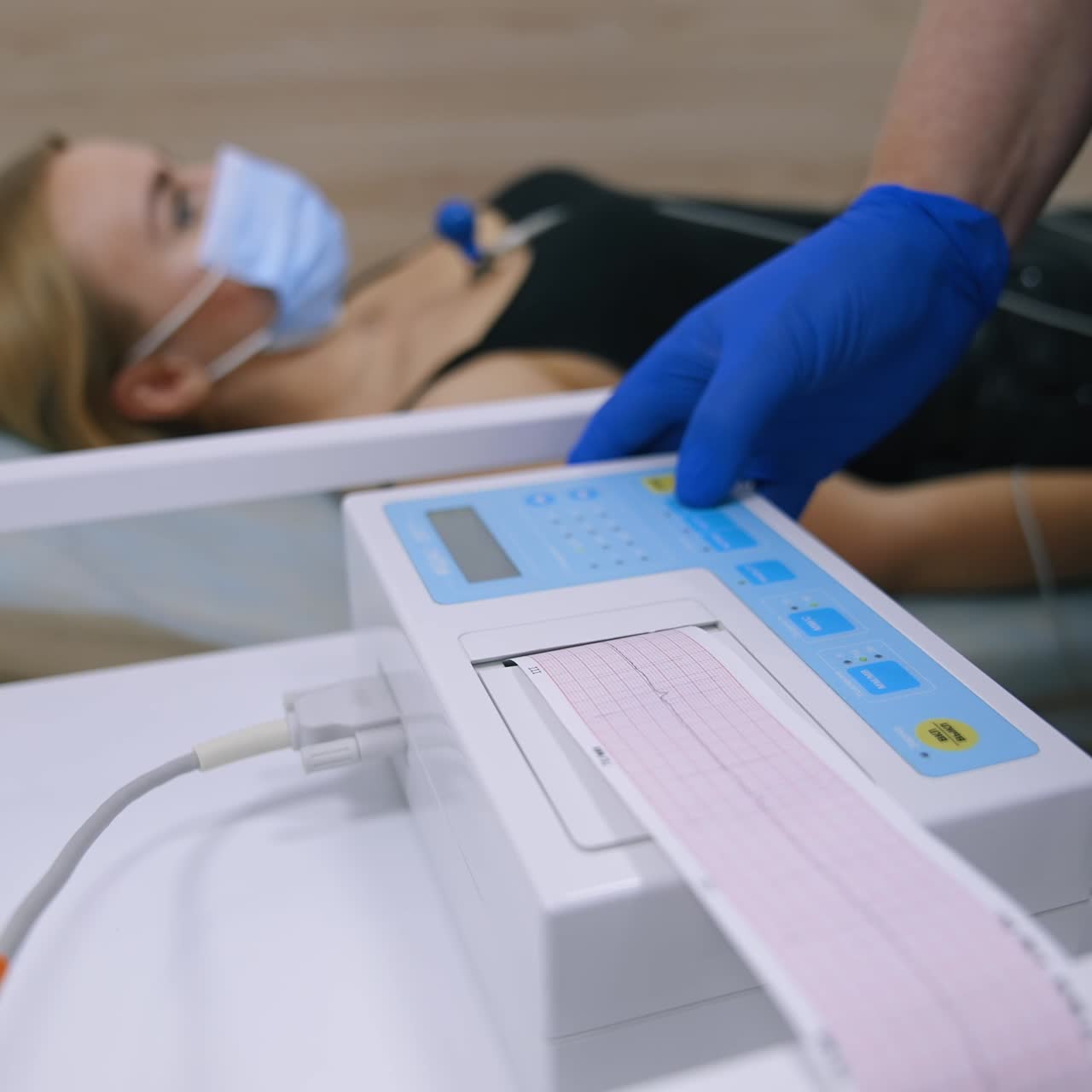 Electrocardiógrafo mostrando el resultado del chequeo cardíaco en papel. Mano de un médico con guantes de látex azules pulsando el botón de la máquina. Paciente tumbado al fondo