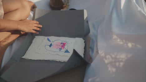 slow motion panning shot to a folder with an abstract painting which is held by the sitting artist on a white linen cloth dressed in a beige dress