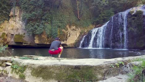 boy sitting alone on the shore and touching the water of a lake in front of a waterfall