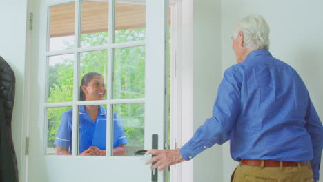 senior man greeting female nurse or care worker making home visit in uniform at door