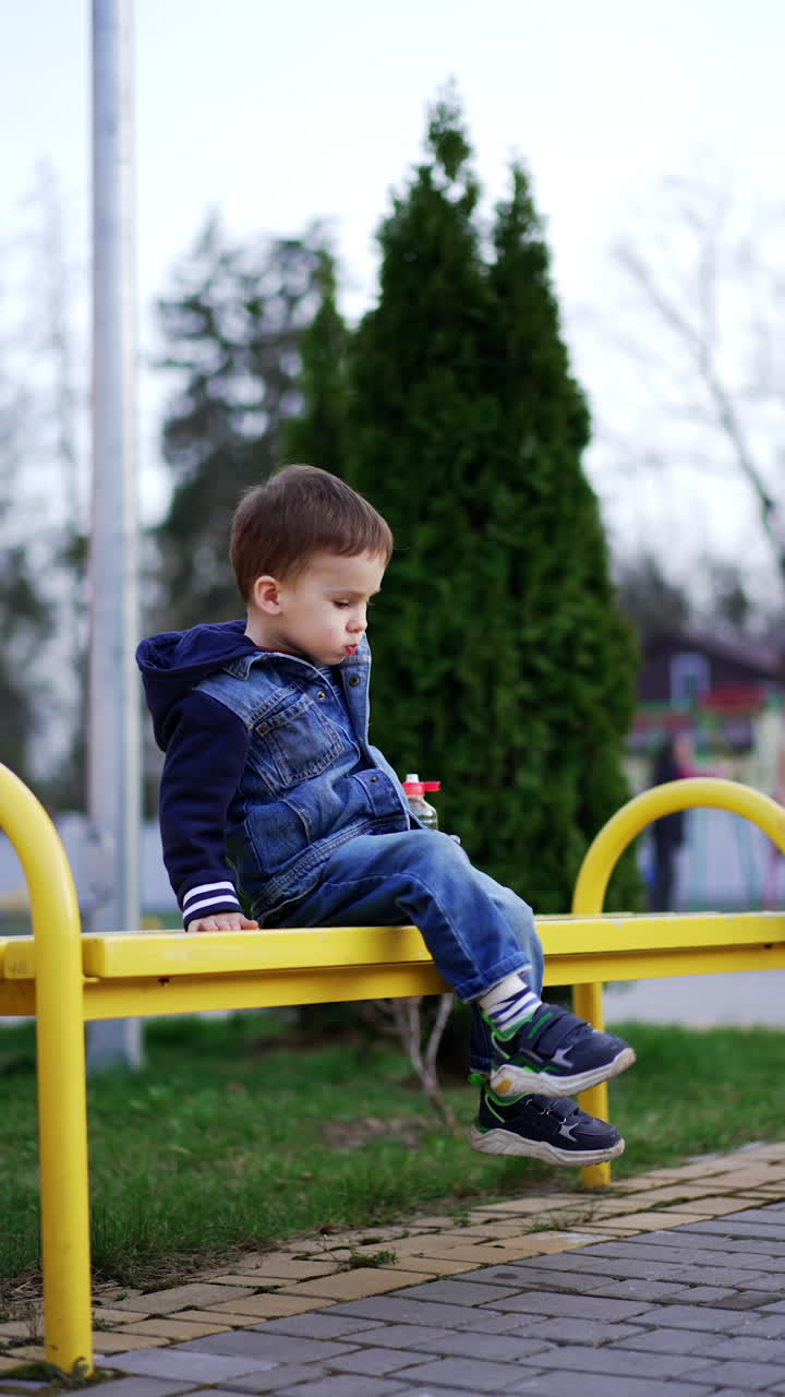 Little Caucasian boy sitting on the bench drinks water from a bottle. Kid stands up and jumps cheerfully. Vertical video.