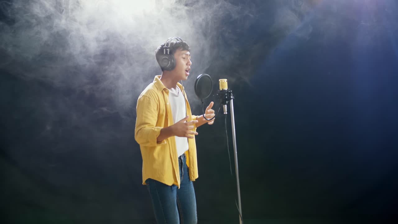 joven asiático con auriculares golpeando en un micrófono condensador en el fondo blanco de humo negro
