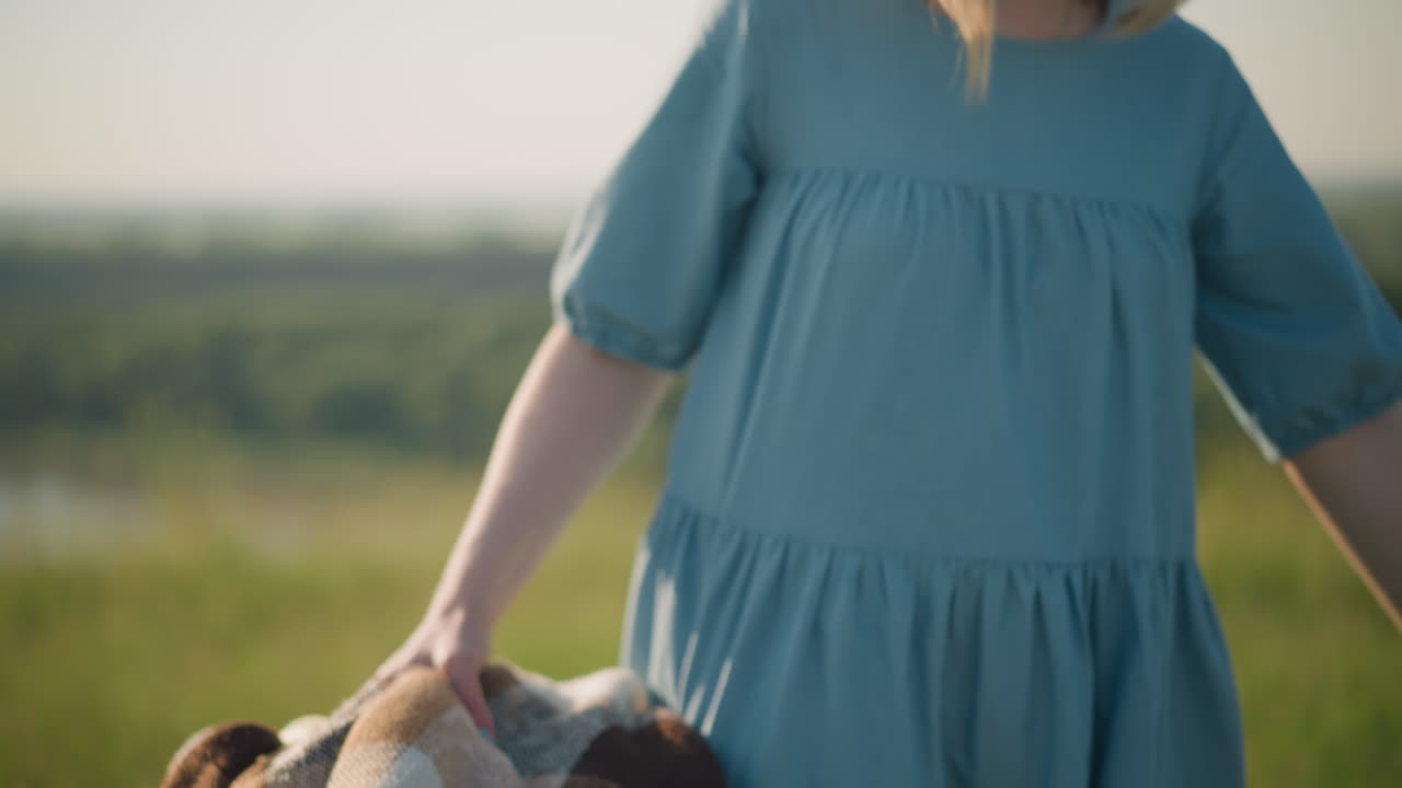 A close-up shot of a mother in a blue dress bending down to pick up a basket filled with fresh fruit from the grass. The scene captures a simple yet warm moment of a picnic in a grassy field