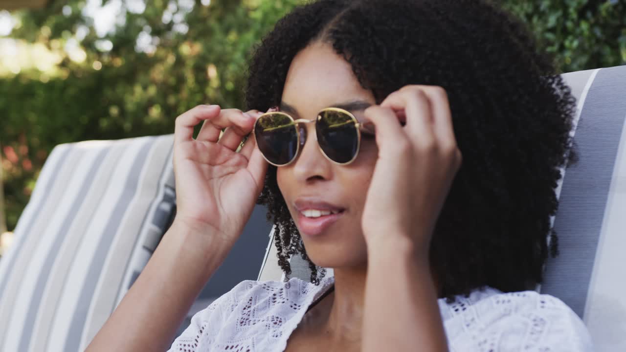 Sous le soleil, une femme afro-américaine en maillot de bain se prélassant au bord de la piscine, mettant ses lunettes de soleil, les ajustant et arrangeant ses cheveux.