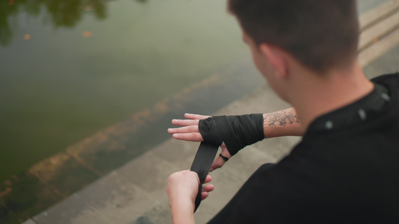 Weißer Mann sichert Handbandagen in der Nähe eines Sees, tätowierte Unterarme, methodische Finger, schwarze Bandage auf Betonstufen, Underground-Kämpfer bereitet sich vor, Trainerinspektion, Wettkämpfer-Routine, rituelles Tapen