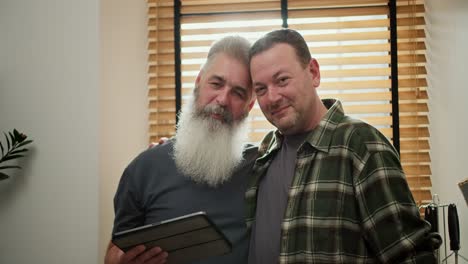 Retrato-De-Una-Feliz-Pareja-De-Dos-Hombres-Homosexuales-Lgbt,-Un-Hombre-De-Mediana-Edad-Con-Cabello-Gris-Y-Barba-Incipiente-Con-Una-Camisa-A-Cuadros-Verde-Junto-Con-Su-Novio-Mayor-Con-Cabello-Gris-Y-Una-Exuberante-Barba-Gris-Con-Una-Camiseta-Gris-Que-Sostiene-Una-Tableta-Electrónica-En-La-Cocina-En-Un-Apartamento-Moderno