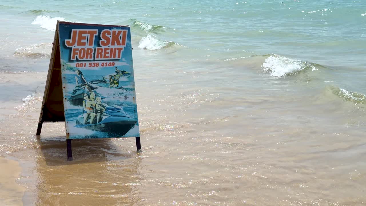 Ein hölzernes A-Rahmen-Jetski-Verleihschild steht an der Küste, während Meereswellen brechen