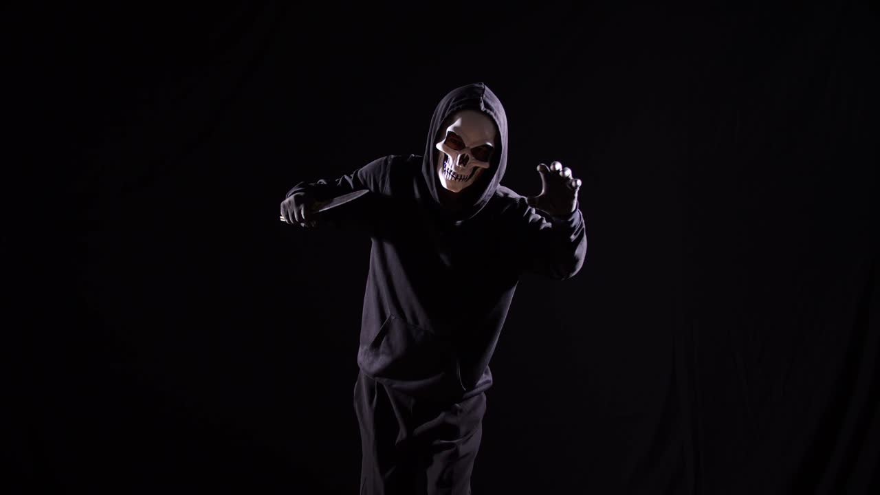 hombre aterrador en la sudadera con capucha con máscara de halloween sosteniendo un cuchillo y haciendo un gesto aterrador mientras camina sobre el fondo negro