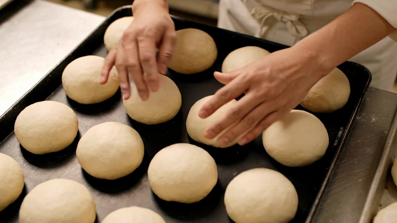 Padeiro com jaqueta e avental pegando e girando bolas de massa, preparando-as para o forno em uma bandeja preta na padaria.