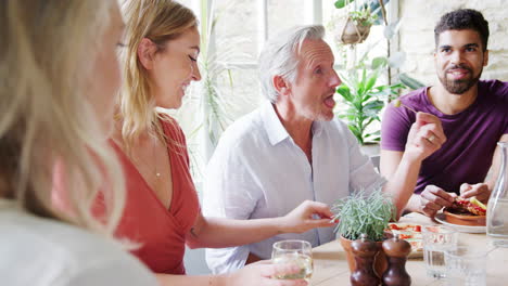 Four-mixed-age-adult-friends-talking-and-eating-lunch-together-at-a-table,-selective-focus