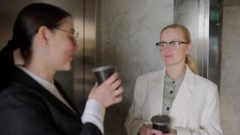 confident brunette girl in round glasses in a business uniform talking with her colleague blonde businesswoman while they are drinking coffee during a break between work in a modern office