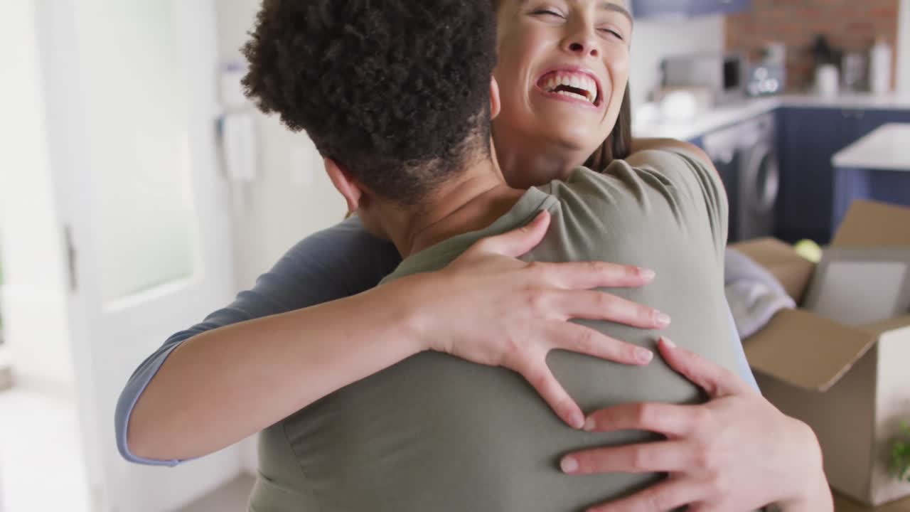 feliz pareja biracial mudándose de casa, abrazándose y sonriendo en la cocina