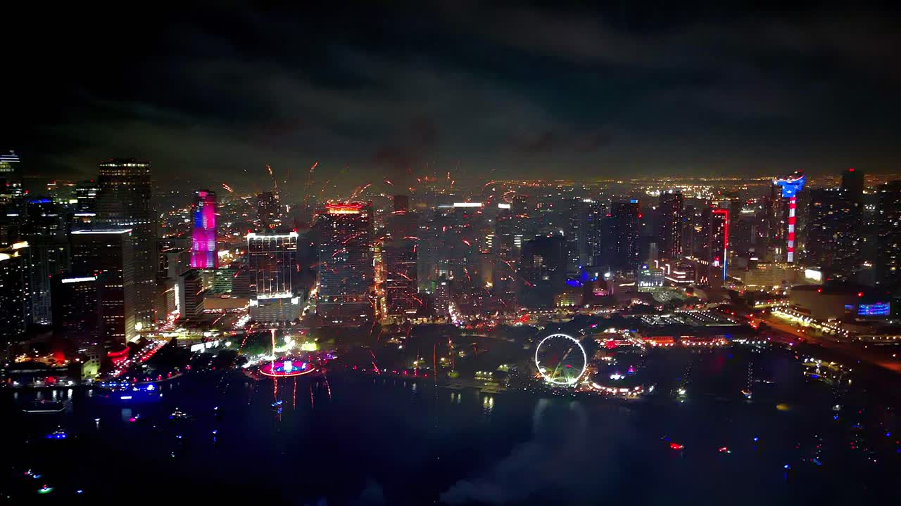 Aerial drone angle showcases fireworks exploding in vivid bursts above the bay, reflecting across the water with radiant brilliance
