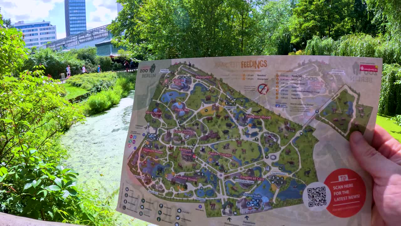 A hand holds a detailed zoo map over a lush, sunlit riverside in Berlin. Camera smoothly pans between the map and the scenic outdoor environment