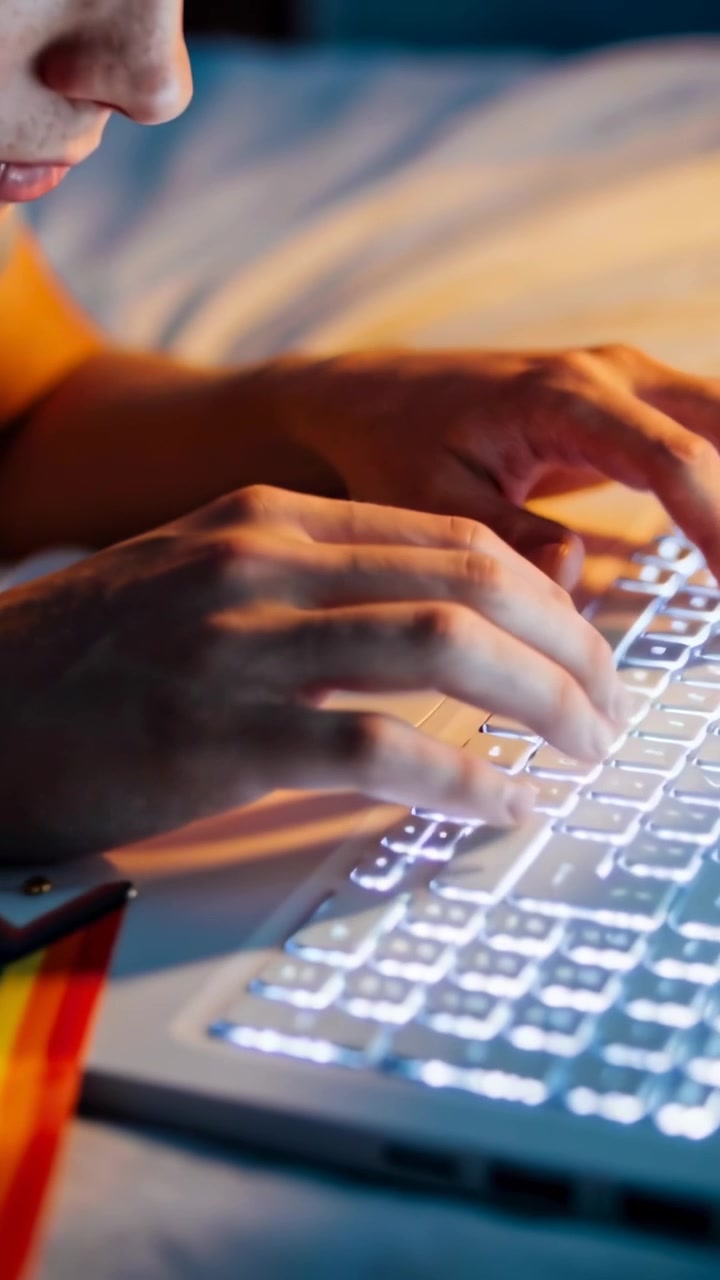 Video verticale: Si vede un uomo con camicia arancione che digita su un laptop retroilluminato sul letto, studia con nastro del Pride