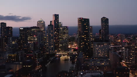 Slide-and-pan-elevated-shot-of-modern-tall-office-and-residential-buildings-in-Canary-Wharf-futuristic-district-in-evening.-City-lights-reflecting-on-water-surface.-London,-UK